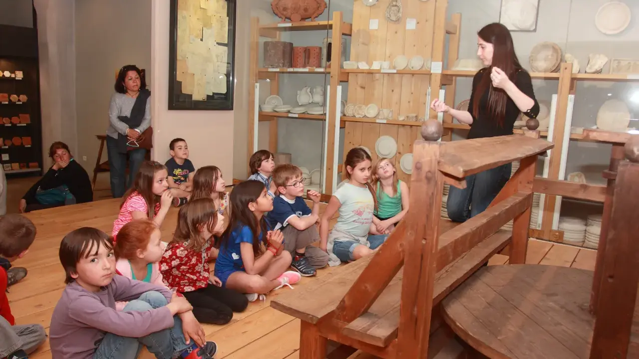 Visite d'un musée à Apt : des enfants écoutent attentivement une guide devant des étagères remplies d'objets anciens.