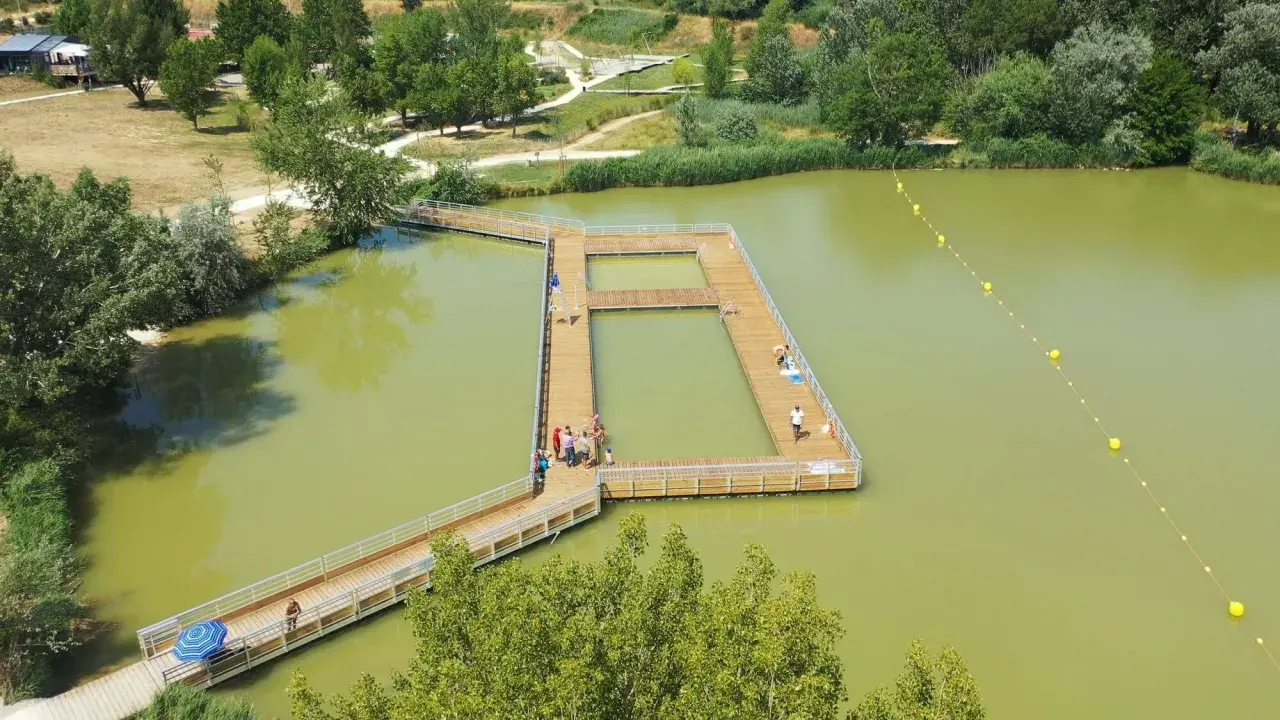 Ponton en bois sur un lac verdâtre, une **base de loisirs** animée avec des baigneurs profitant du soleil.