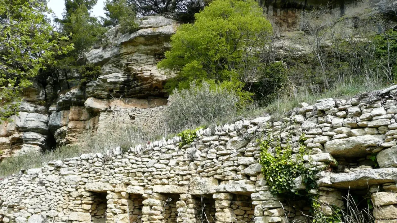Vestiges d'une ancienne ville de murs en pierre, intégrée à une falaise rocheuse, avec végétation luxuriante.