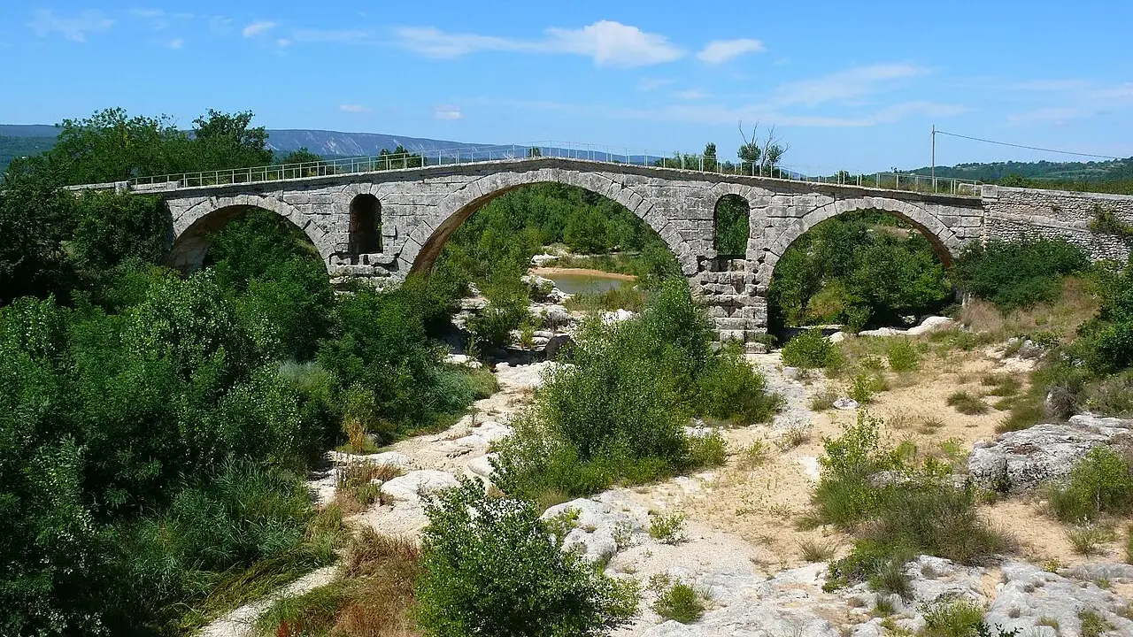 Vieux pont de pierre à arches sur une rivière asséchée, entouré de végétation luxuriante.