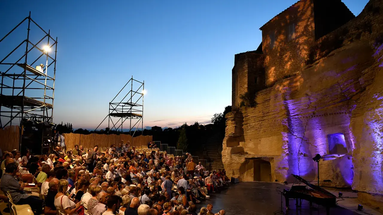 Public venu pour le festival de Gordes, une foule assise attend un concert au crépuscule, avec un piano sur scène et des structures métalliques.