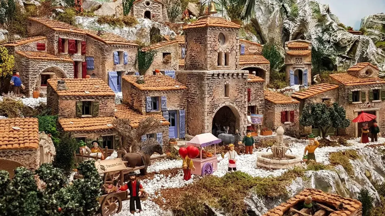 Une **crèche provençale** animée avec des santons, des maisons en pierre, un âne, un chariot et une fontaine, le tout dans un paysage rocheux.