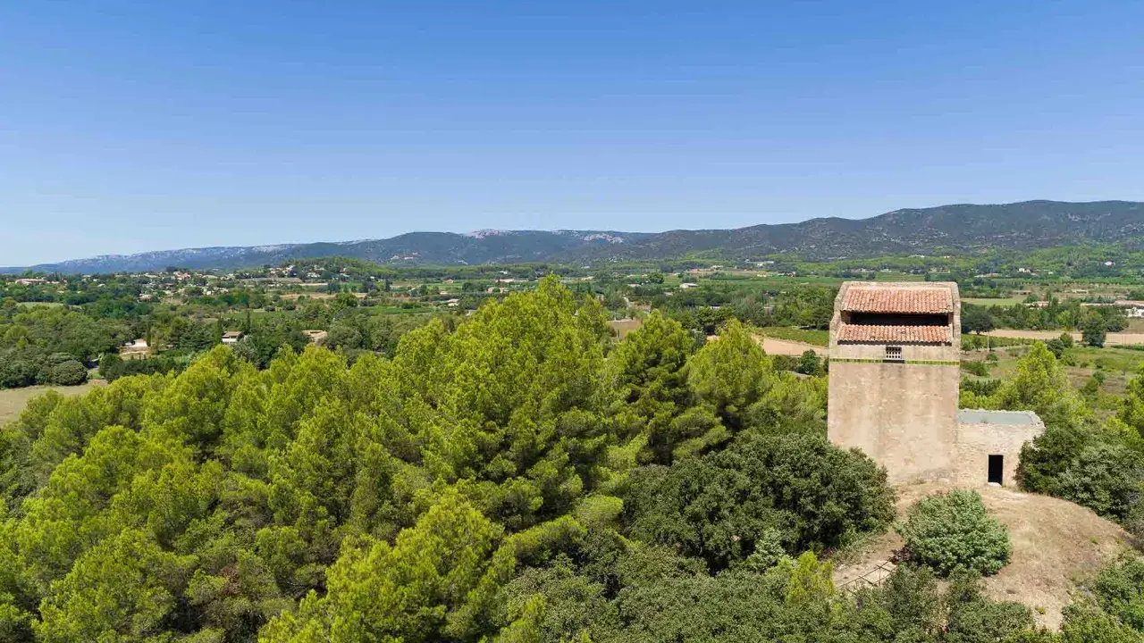 Vue panoramique sur la campagne provençale avec le moulin de Puyvert, entouré de pins et de verdure, sous un ciel bleu éclatant.