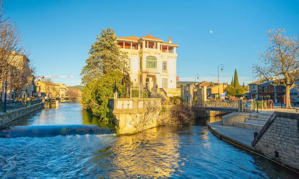 L'eau vive de la Sorgue traverse L'Isle-sur-la-Sorgue, bordée de maisons et d'un bâtiment blanc imposant.