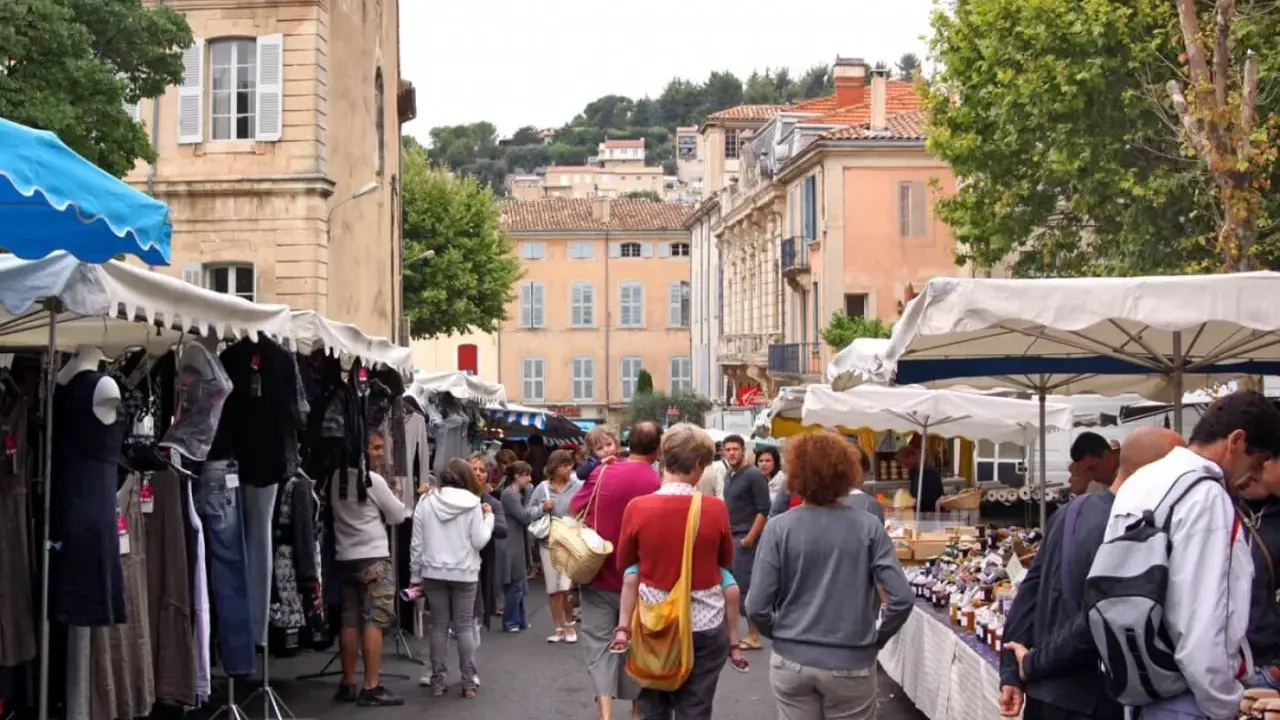 Ambiance animée au marché d'Apt, avec des étals de vêtements et de produits locaux. Des passants flânent sous les parasols, profitant de l'atmosphère provençale.