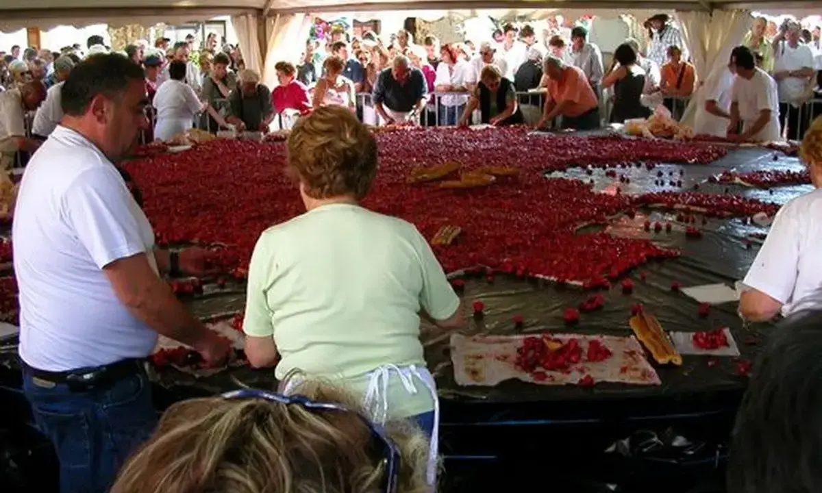 Des gens préparent une immense tarte aux fraises pour la fête de la fraise. Des fraises fraîches et des morceaux de pain sont disposés sur une grande table.