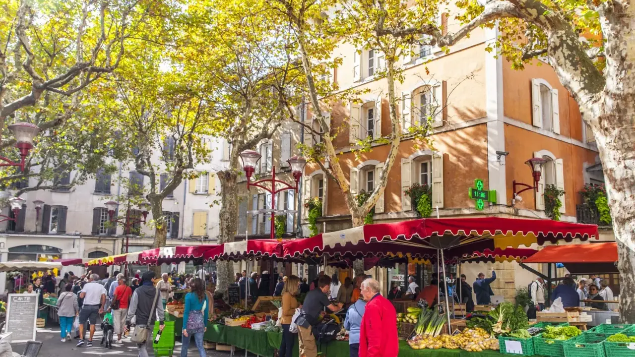 Ambiance animée au marché de Manosque, sous les platanes aux feuilles d'automne. Étals colorés de fruits et légumes frais, passants flânant.