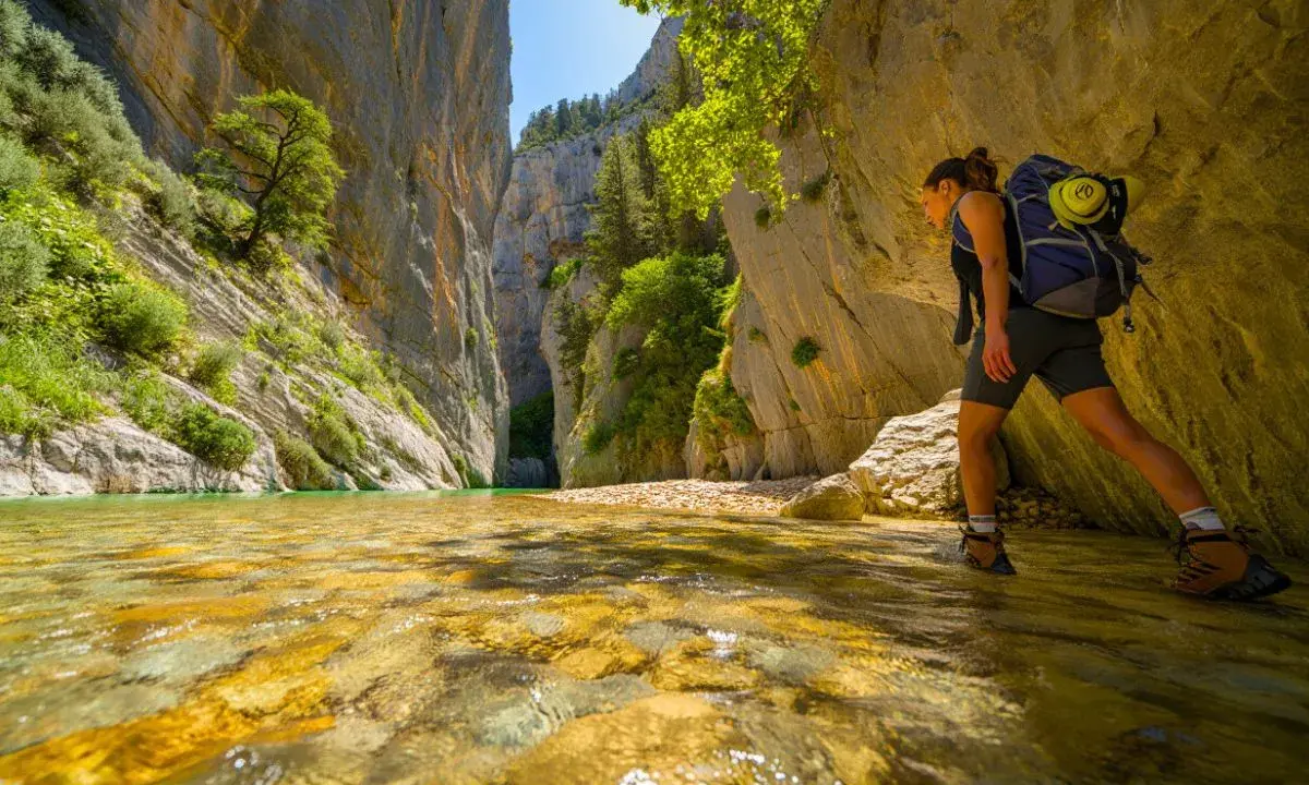 Randonneuse explorant la gorge du Toulourenc, traversant une rivière aux eaux cristallines bordée de falaises imposantes et de végétation luxuriante.