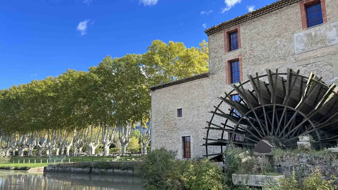 Vieux moulin en pierre avec sa roue à aubes, bordé par une rivière et une allée d'arbres aux couleurs d'automne. Les taillades semblent figées dans le temps.