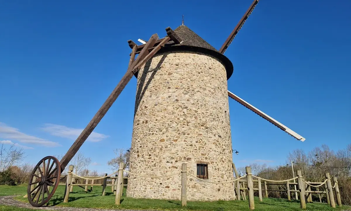 Le moulin de Maryline, une tour de pierre imposante sous un ciel bleu, se dresse fièrement avec ses ailes en bois.