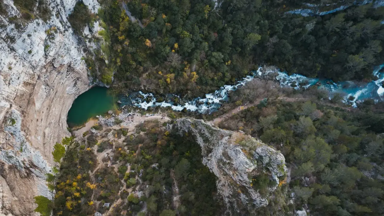 Vue aérienne de la Fontaine de Vaucluse, avec son bassin émeraude et la rivière Sorgue serpentant à travers une gorge boisée.
