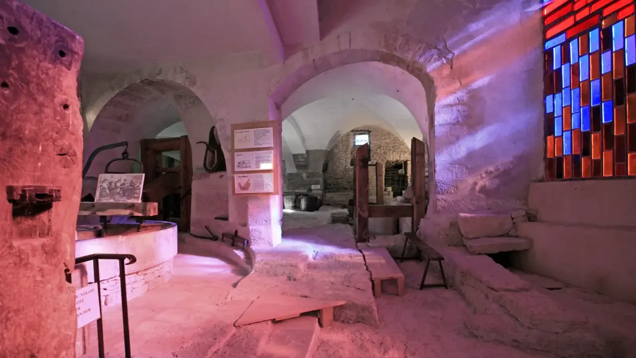 Intérieur d'un musée du verre et du vitrail, avec des arches en pierre, des outils anciens et un vitrail coloré projetant des rayons lumineux.