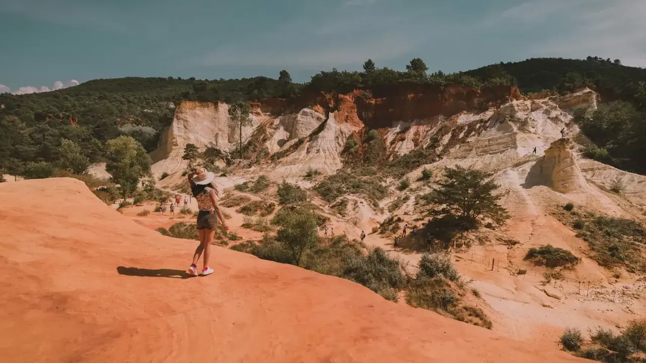 Une femme au chapeau admire le paysage spectaculaire de la carrière d'ocre du Vaucluse, avec ses falaises colorées et sa végétation luxuriante.