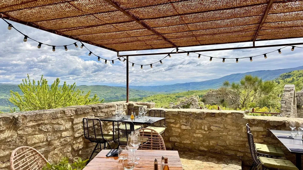 Terrasse d'un restaurant avec vue panoramique sur les collines. Tables dressées sous un auvent de chaume, ambiance estivale. Un jardin sur le toit Saignon promet un repas inoubliable.