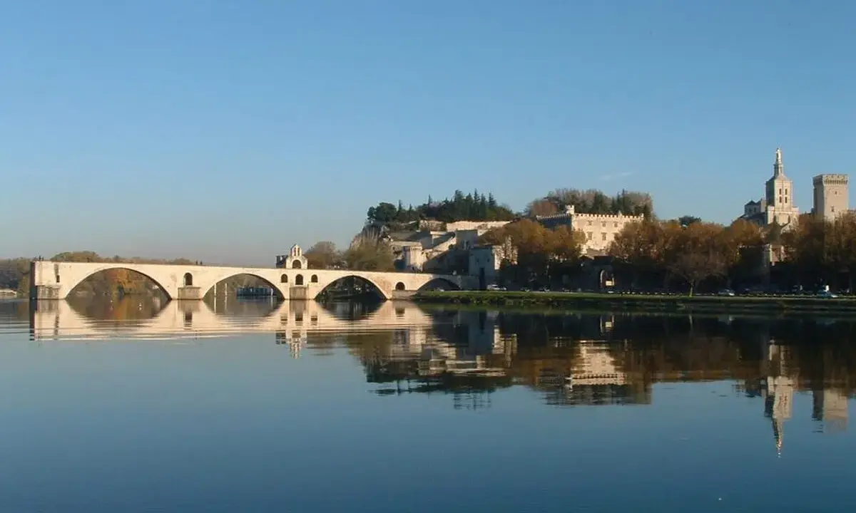 Le pont d'Avignon se reflète dans le Rhône, avec le Palais des Papes et l'île de la Barthelasse en arrière-plan.