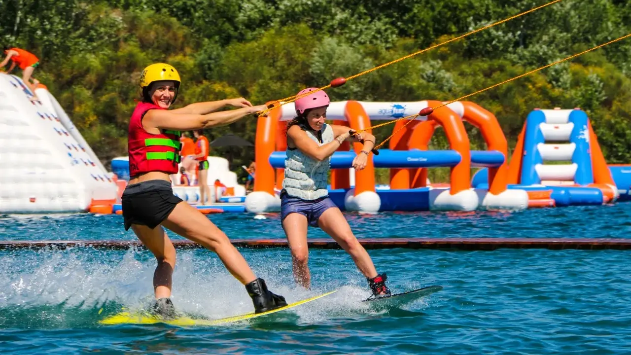 Deux femmes font du wakeboard dans un wam park orange et bleu, avec des structures gonflables en arrière-plan.