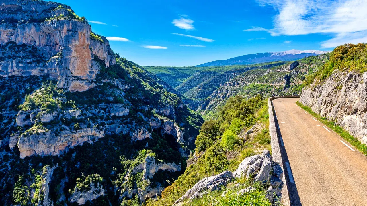 Route sinueuse longeant les impressionnantes gorges de la Nesque, avec des falaises rocheuses et une végétation luxuriante sous un ciel bleu.