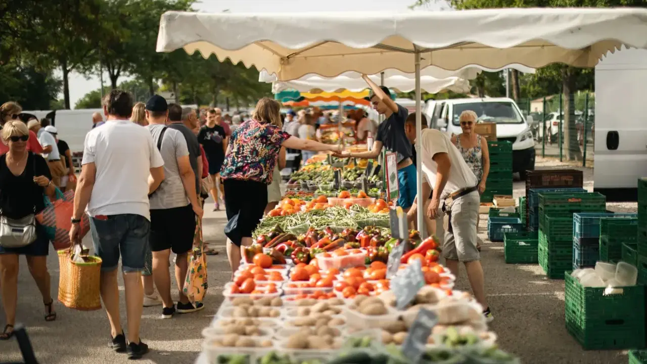 Un marché animé, les angles des étals débordent de fruits et légumes frais. Des clients font leurs courses sous les parasols blancs.