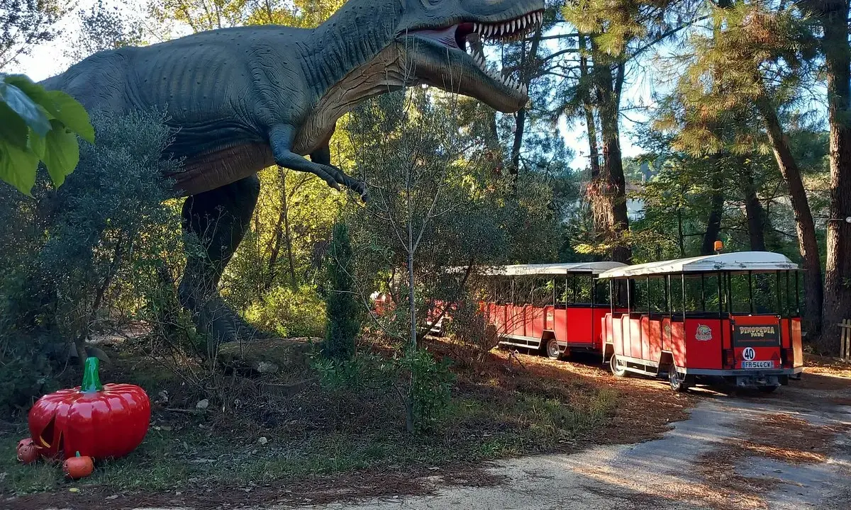 Un train rouge traverse le parc Grimmland, passant près d'un dinosaure géant et de citrouilles décoratives.