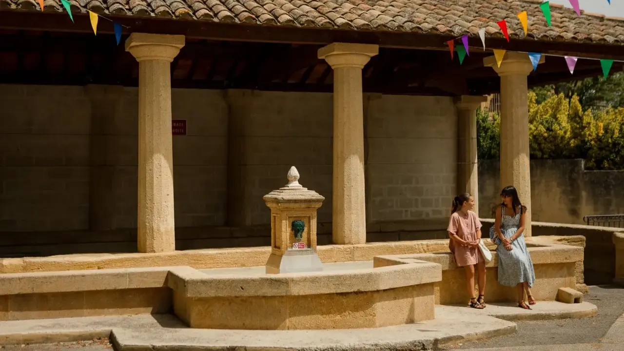 Deux femmes discutent près d'une fontaine sous un auvent orné de fanions colorés, ambiance de marché à Rochefort-du-Gard.