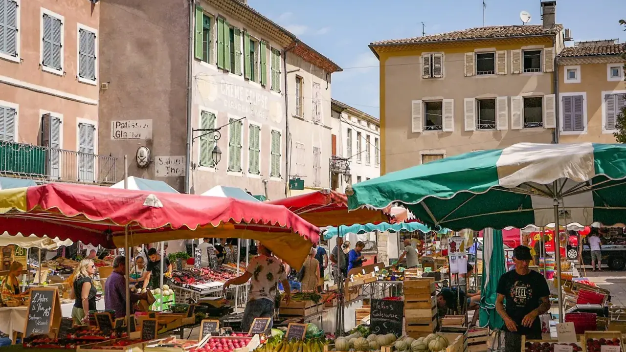 Le marché le Thor animé, avec des étals colorés de fruits et légumes sous des parasols. Des passants flânent entre les stands, profitant du soleil provençal.