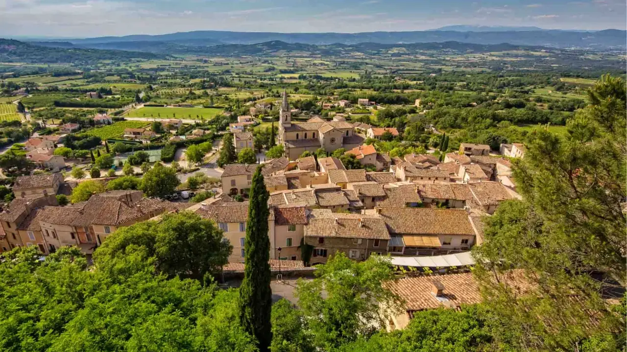 Vue aérienne du village de Bonnieux, avec ses toits de tuiles ocres, son église et la campagne environnante verdoyante. Le marché de Bonnieux est un lieu animé.