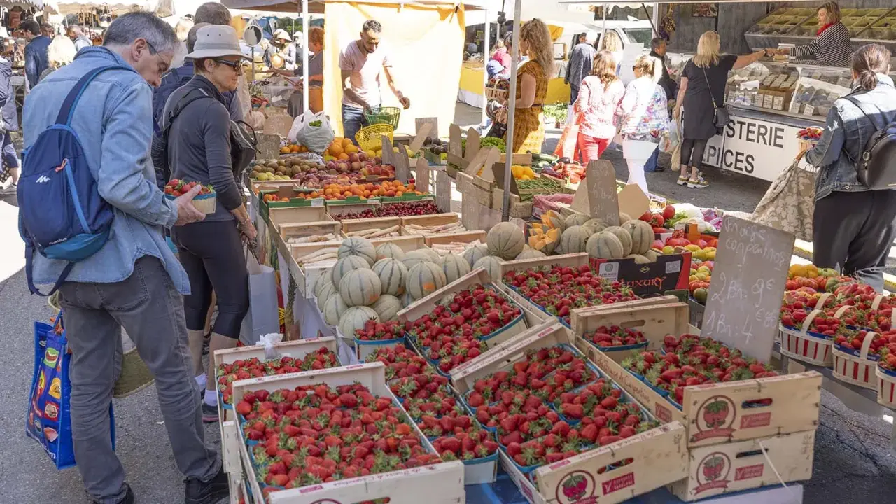 Le marché de Forcalquier animé, avec des étals débordant de fraises, melons et fruits. Des clients font leurs achats sous le soleil.