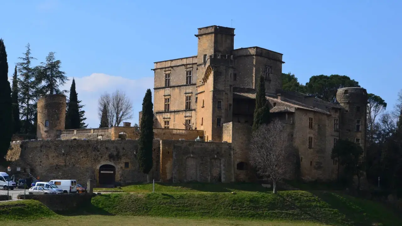 Le majestueux **Château de Lourmarin** se dresse sur une colline, ses pierres ocres baignées de soleil sous un ciel bleu. Des cyprès élancés l'entourent.
