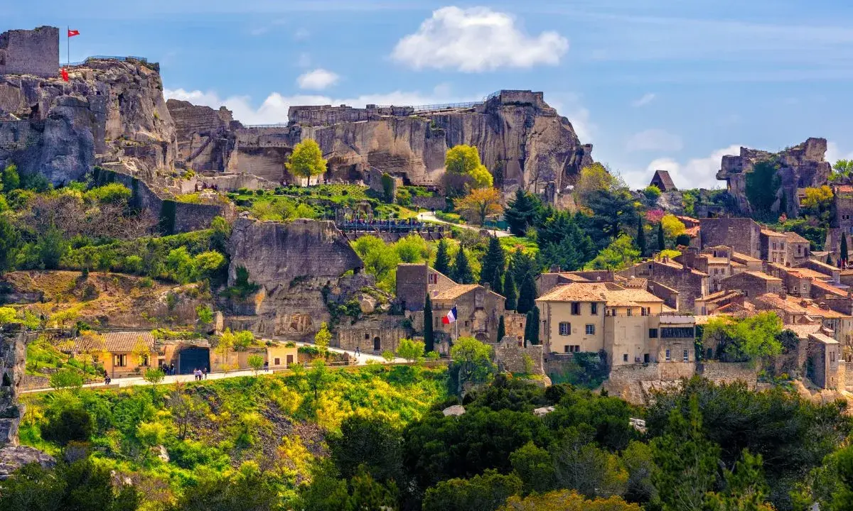 Vue panoramique de la route des villages perchés, avec un château en ruines dominant des maisons en pierre et une végétation luxuriante sous un ciel bleu.