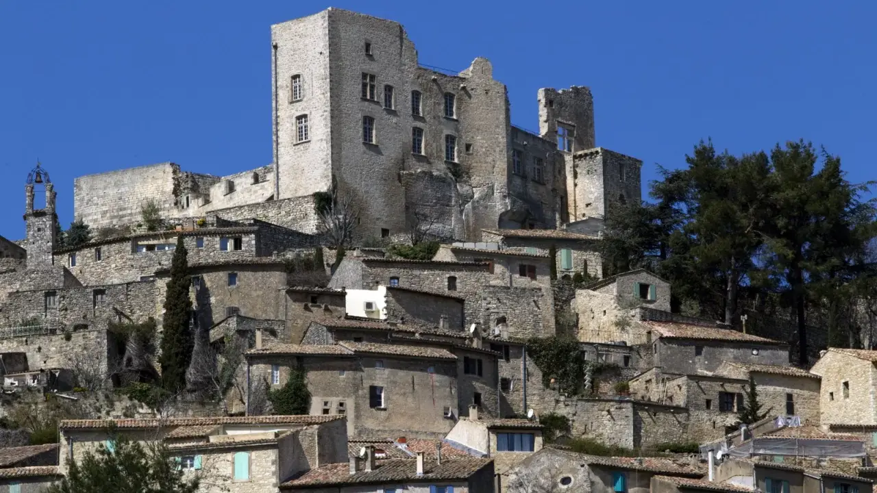 Vue d'un village perché dominé par les ruines imposantes du **château du marquis de Sade**, sous un ciel bleu éclatant.
