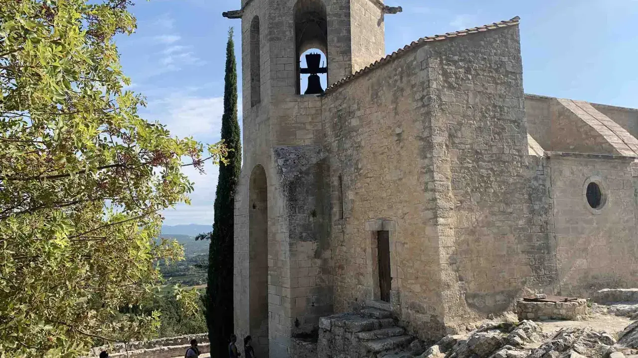 Le clocher de Notre-Dame d'Alidon, avec sa cloche visible, se dresse fièrement sous un ciel bleu. Des visiteurs admirent le paysage.