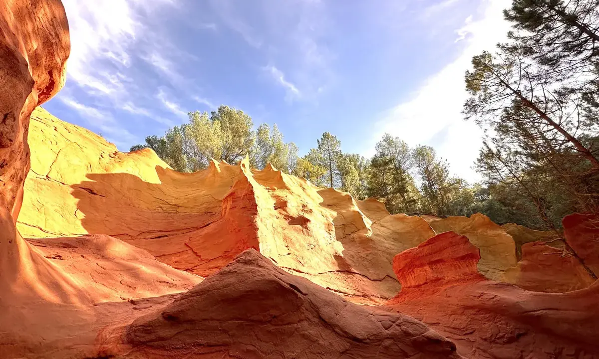 Carrières d'ocre aux couleurs vives, rappelant l'usine d'ocre Mathieu. Le ciel bleu et les arbres verts contrastent avec les roches rouges et jaunes.
