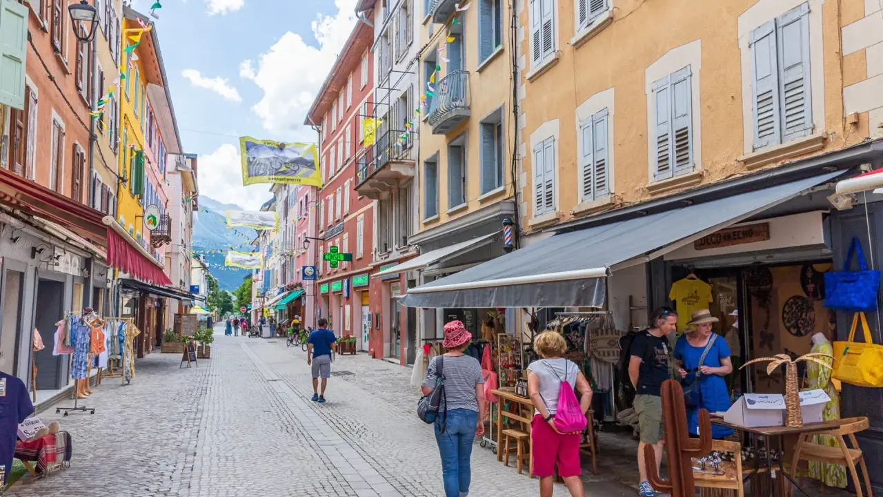 Rue commerçante animée à Aubenas-les-Alpes, avec des façades colorées, des drapeaux et des passants profitant de l'été.