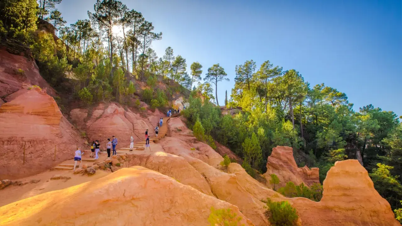 Des randonneurs explorent un sentier boisé aux ocres de Provence, baigné de soleil.