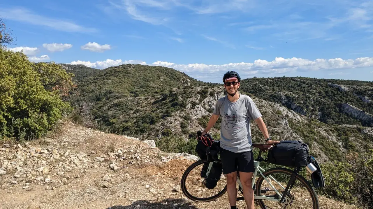 Cycliste souriant avec son vélo chargé, profitant d'une pause lors d'un tour du Luberon à vélo. Paysage rocailleux et collines verdoyantes sous un ciel bleu.