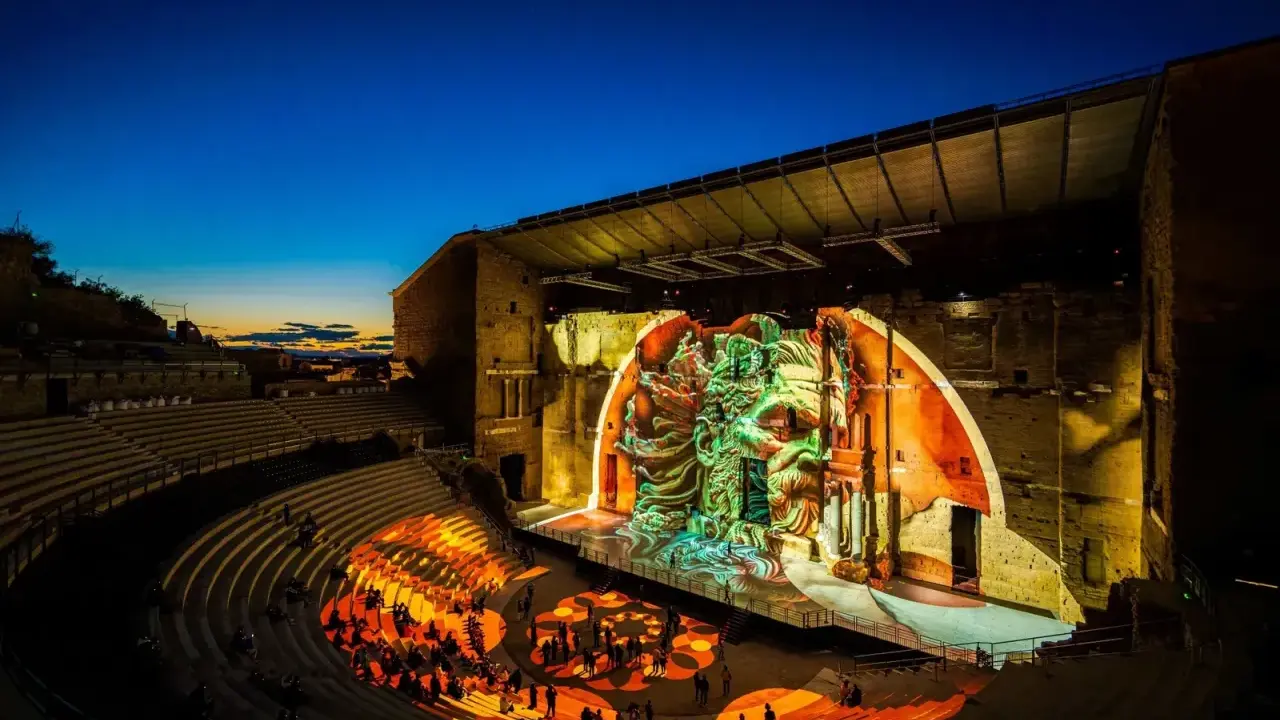 Spectacle son et lumière dans un théâtre antique. Le parvis orange illumine les gradins où le public est assis, tandis que la scène projette une fresque monumentale.