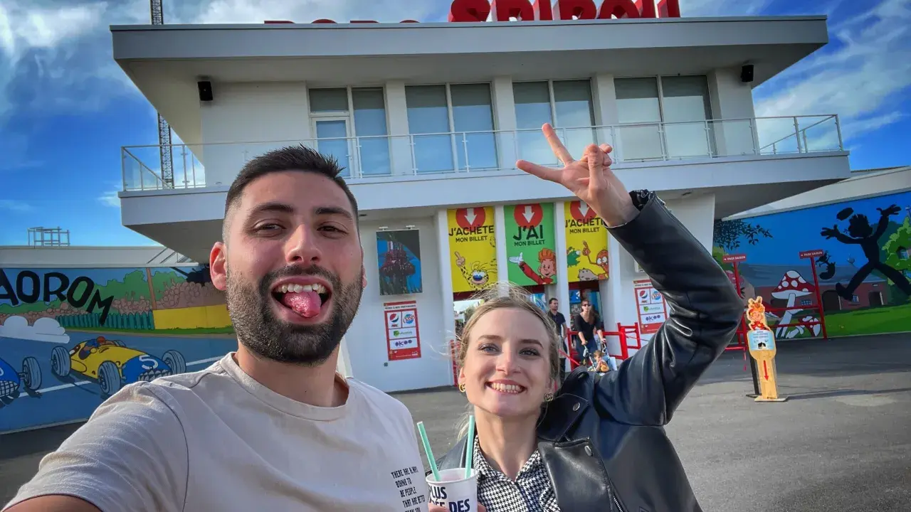 Un couple s'amuse devant le parc Spirou. La femme fait un signe de paix. Combien de temps pour faire le parc Spirou ? Une journée entière !