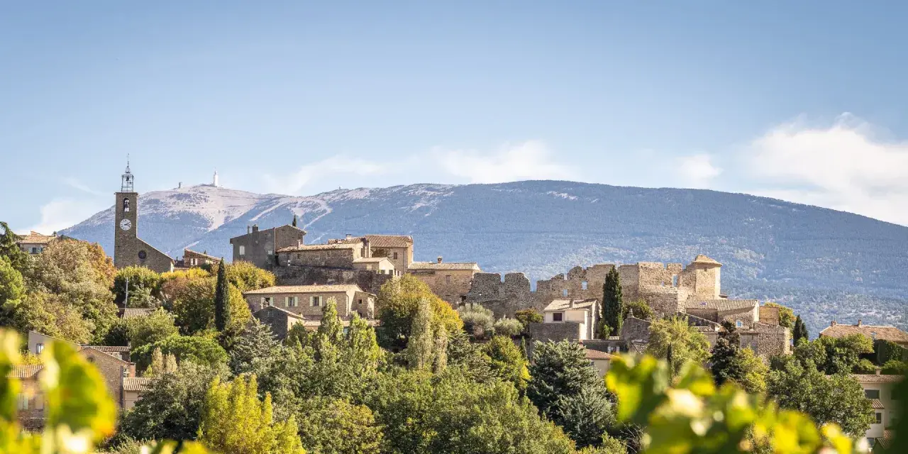 Vue d'un faucon village perché sur une colline, avec une église et des ruines médiévales. Le Mont Ventoux domine l'horizon.