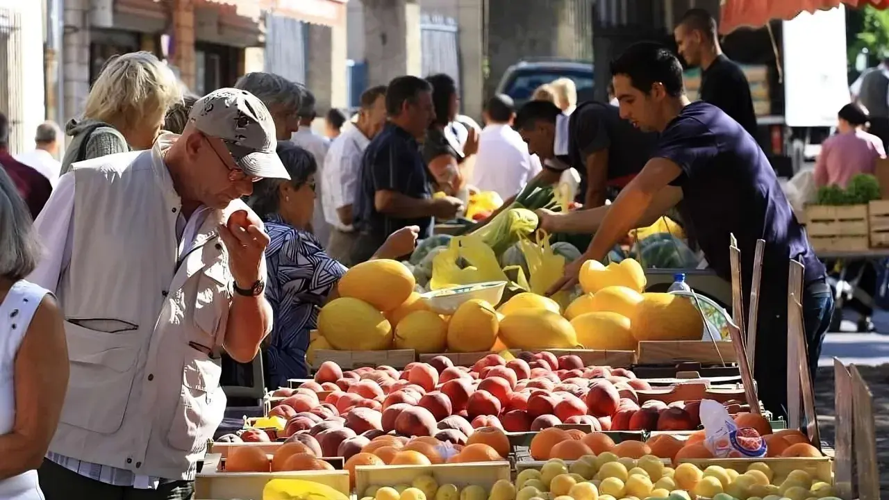 Ambiance animée au marché de Pernes-les-Fontaines. Des étals débordent de fruits colorés, pêches, melons, et agrumes. Des clients choisissent leurs produits.