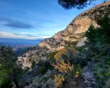 Vue panoramique des gorges de Badarel, avec un sentier rocailleux serpentant à travers la végétation méditerranéenne et une falaise imposante baignée de soleil.