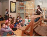 Visite d'un musée à Apt : des enfants écoutent attentivement une guide devant des étagères remplies d'objets anciens.
