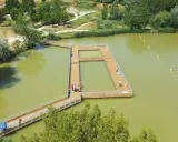 Ponton en bois sur un lac verdâtre, une **base de loisirs** animée avec des baigneurs profitant du soleil.