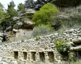 Vestiges d'une ancienne ville de murs en pierre, intégrée à une falaise rocheuse, avec végétation luxuriante.