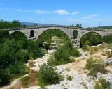 Vieux pont de pierre à arches sur une rivière asséchée, entouré de végétation luxuriante.