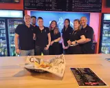 L'équipe du bar Elyzion, souriante, pose devant les réfrigérateurs. Sur le comptoir, des nachos et un menu.