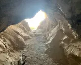 L'entrée de la grotte de Saint Eucher s'ouvre sur une lumière vive, révélant des parois rocheuses sculptées par le temps. Un morceau de bois mort repose au sol.