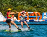 Deux femmes font du wakeboard dans un wam park orange et bleu, avec des structures gonflables en arrière-plan.