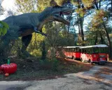 Un train rouge traverse le parc Grimmland, passant près d'un dinosaure géant et de citrouilles décoratives.