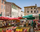 Le marché le Thor animé, avec des étals colorés de fruits et légumes sous des parasols. Des passants flânent entre les stands, profitant du soleil provençal.