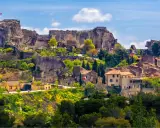 Vue panoramique de la route des villages perchés, avec un château en ruines dominant des maisons en pierre et une végétation luxuriante sous un ciel bleu.